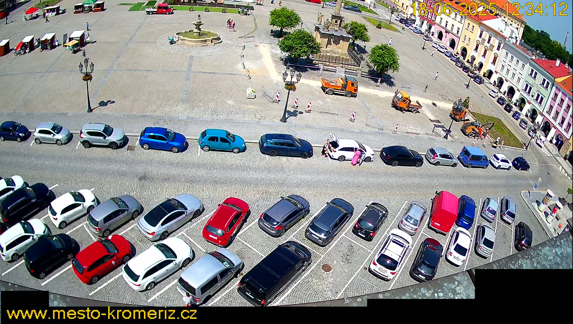 A sunny town square with parked cars, construction equipment, and a fountain in the center.