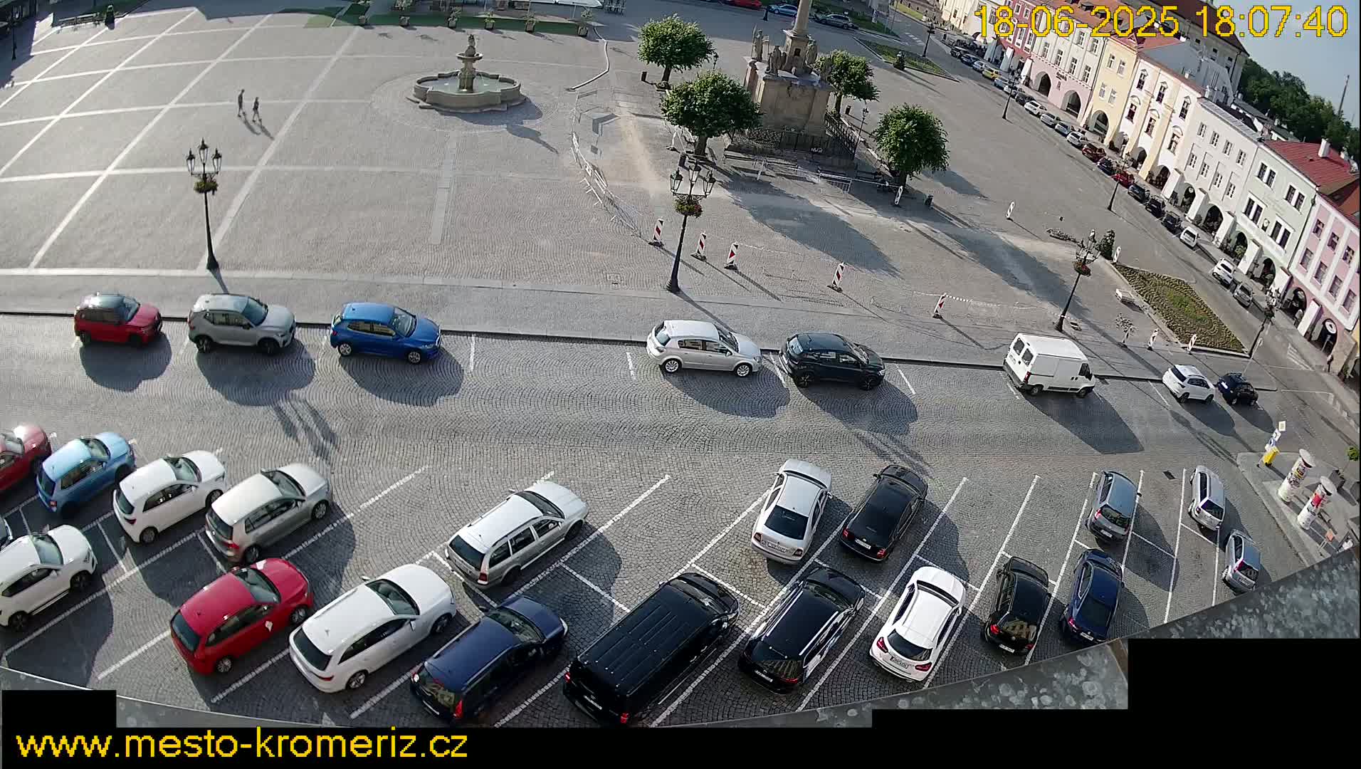A town square with a fountain and several cars parked in a lot, viewed from above on a sunny day.