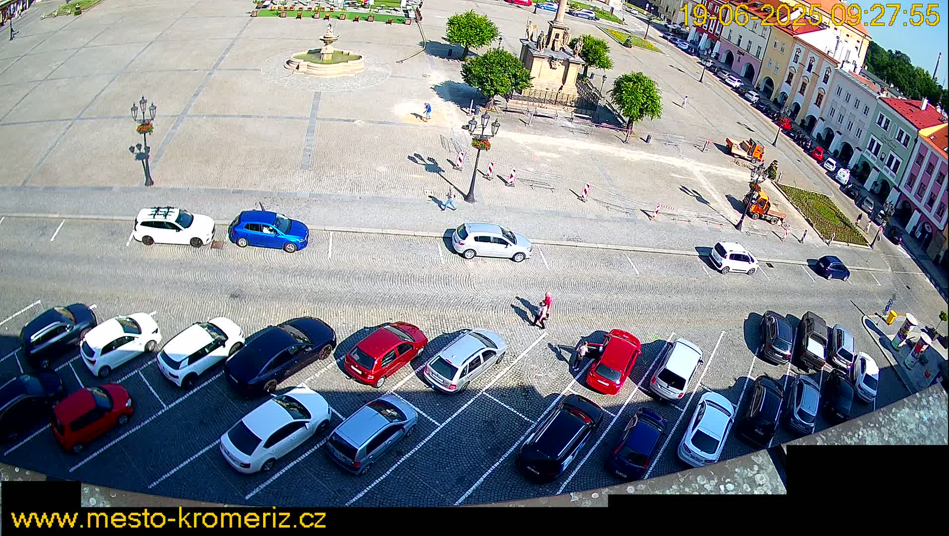 A sunny day shows a town square with a fountain and several cars parked along the side in a parking lot, while some construction or maintenance is happening in the center of the square.