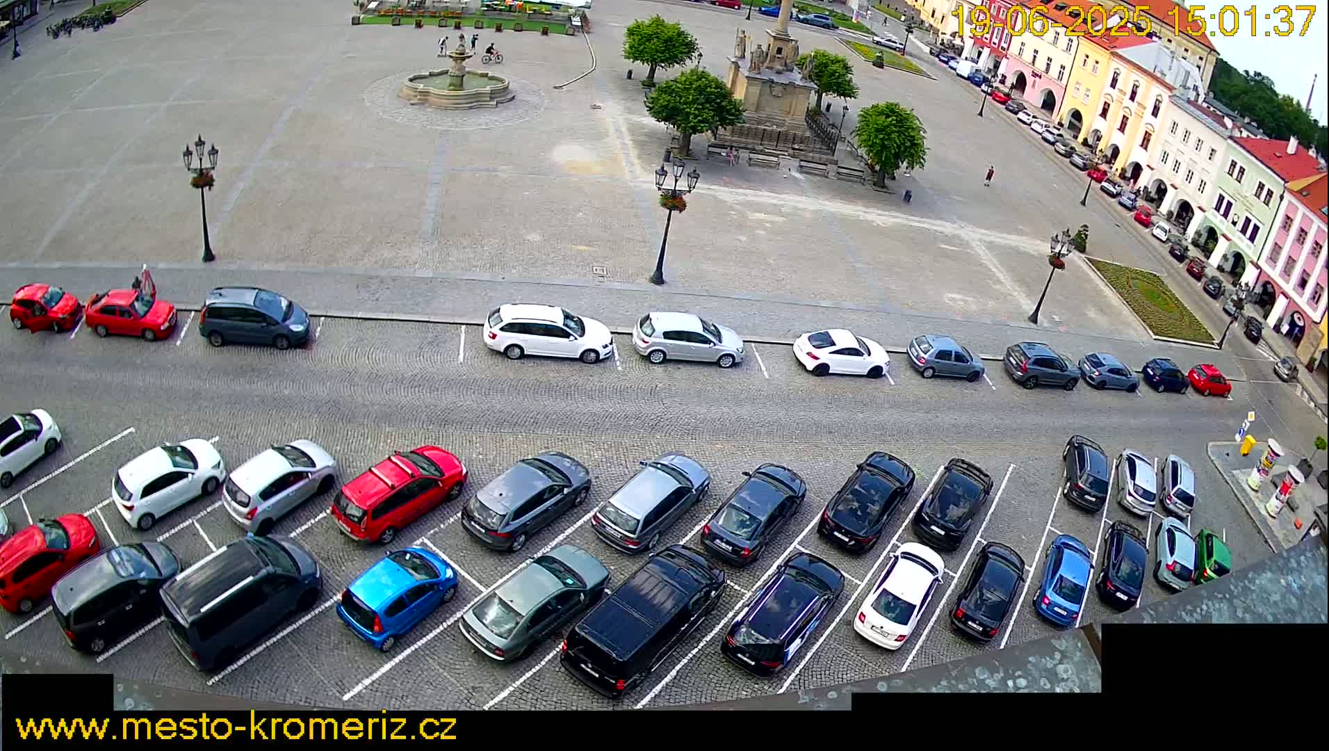 A town square with a fountain and several parked cars under a partly sunny sky.