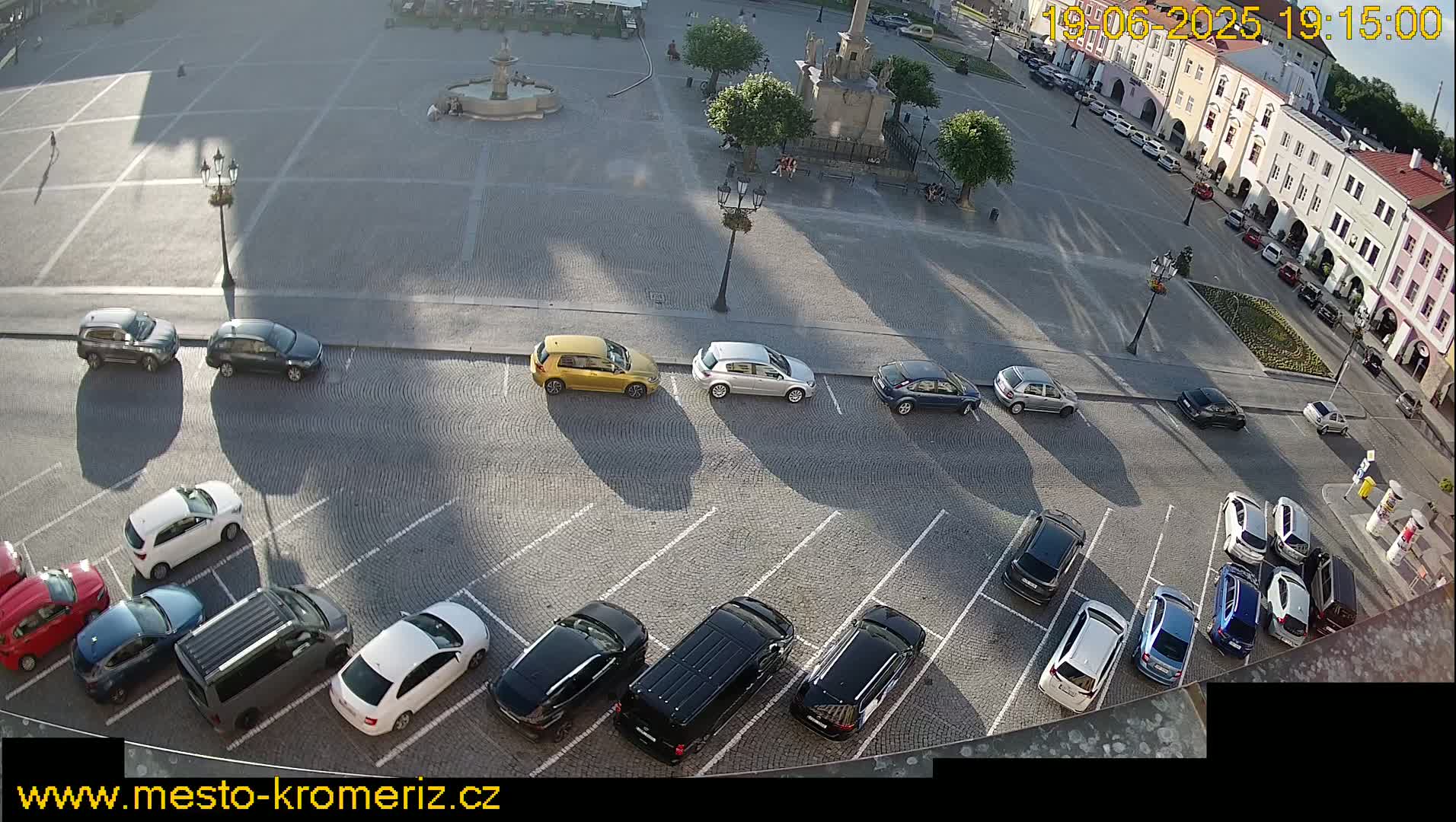 A sunny town square with cars parked in a lot next to it.
