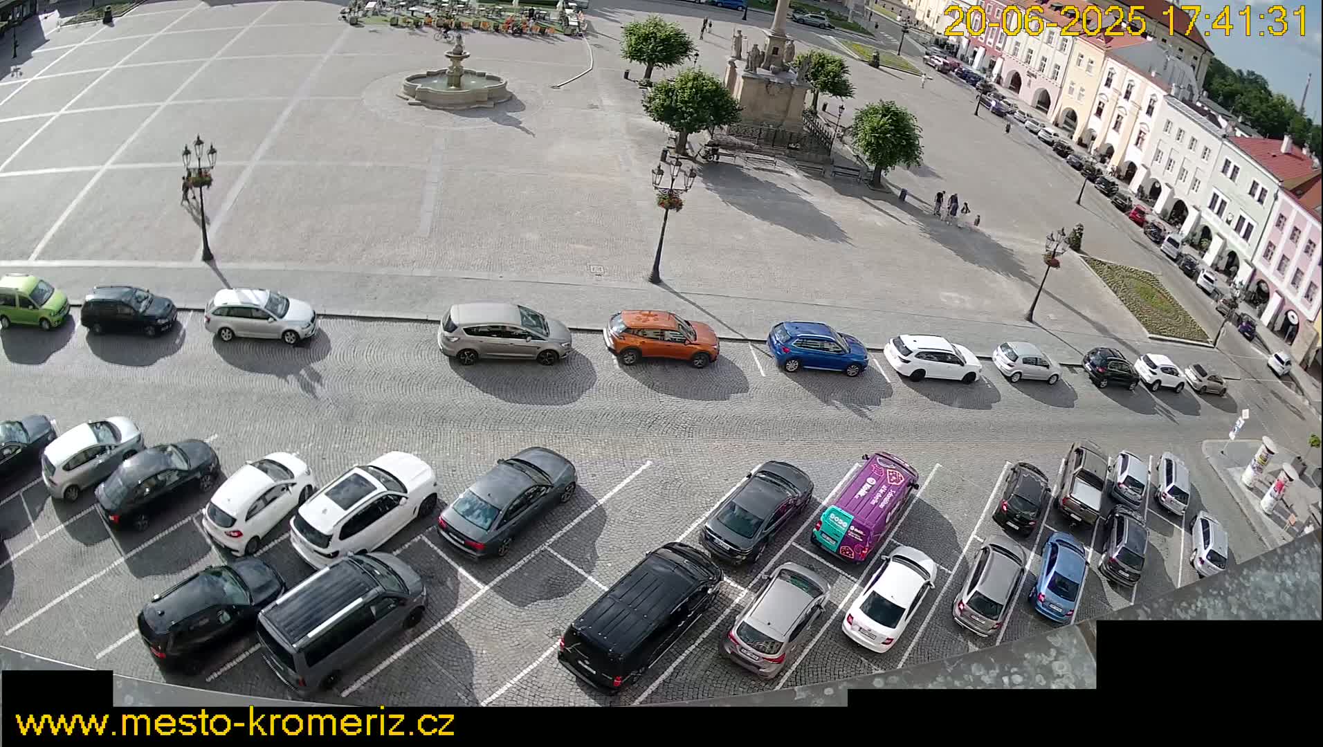 A town square with a fountain and several cars parked along the sides, under sunny conditions.