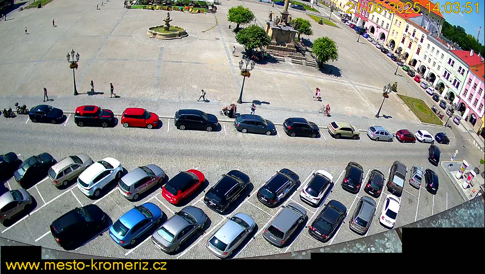 A sunny day shows a town square with a fountain, several parked cars, and a few pedestrians.