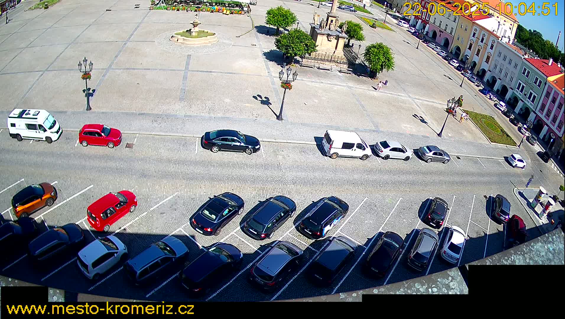 A high-angle view of a town square on a sunny day, with cars parked in a lot surrounding a cobblestone plaza with a fountain.
