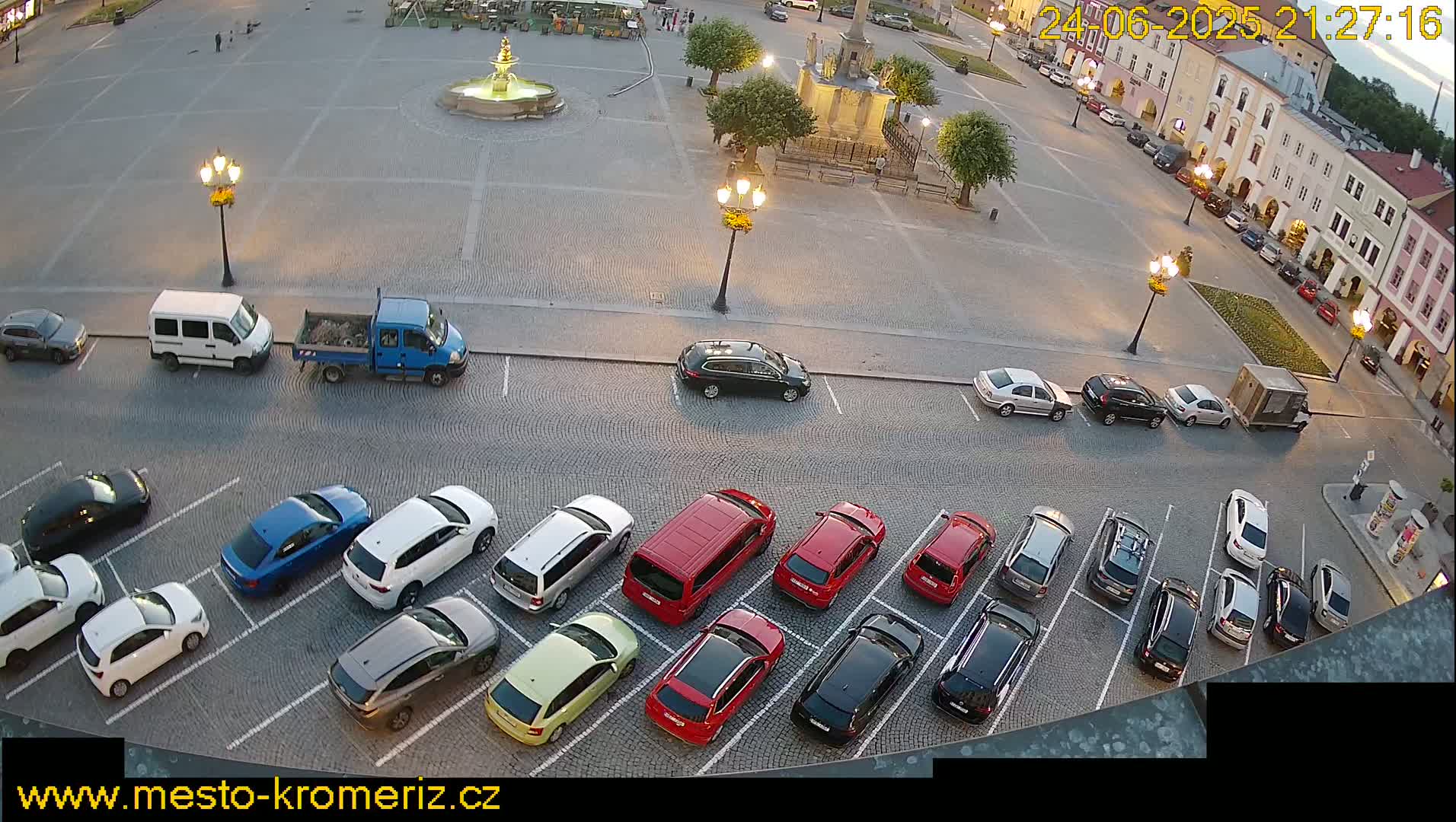 An aerial view of a town square at dusk, showing several cars parked in a lot alongside buildings and a fountain.
