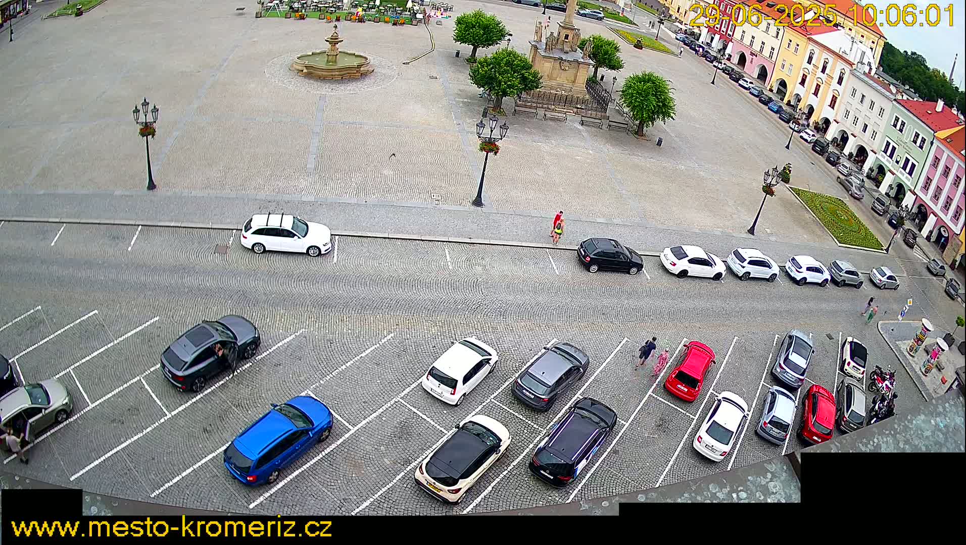 A cobblestone town square with a fountain and surrounding buildings, and a parking lot filled with cars is viewed from above on an overcast day.