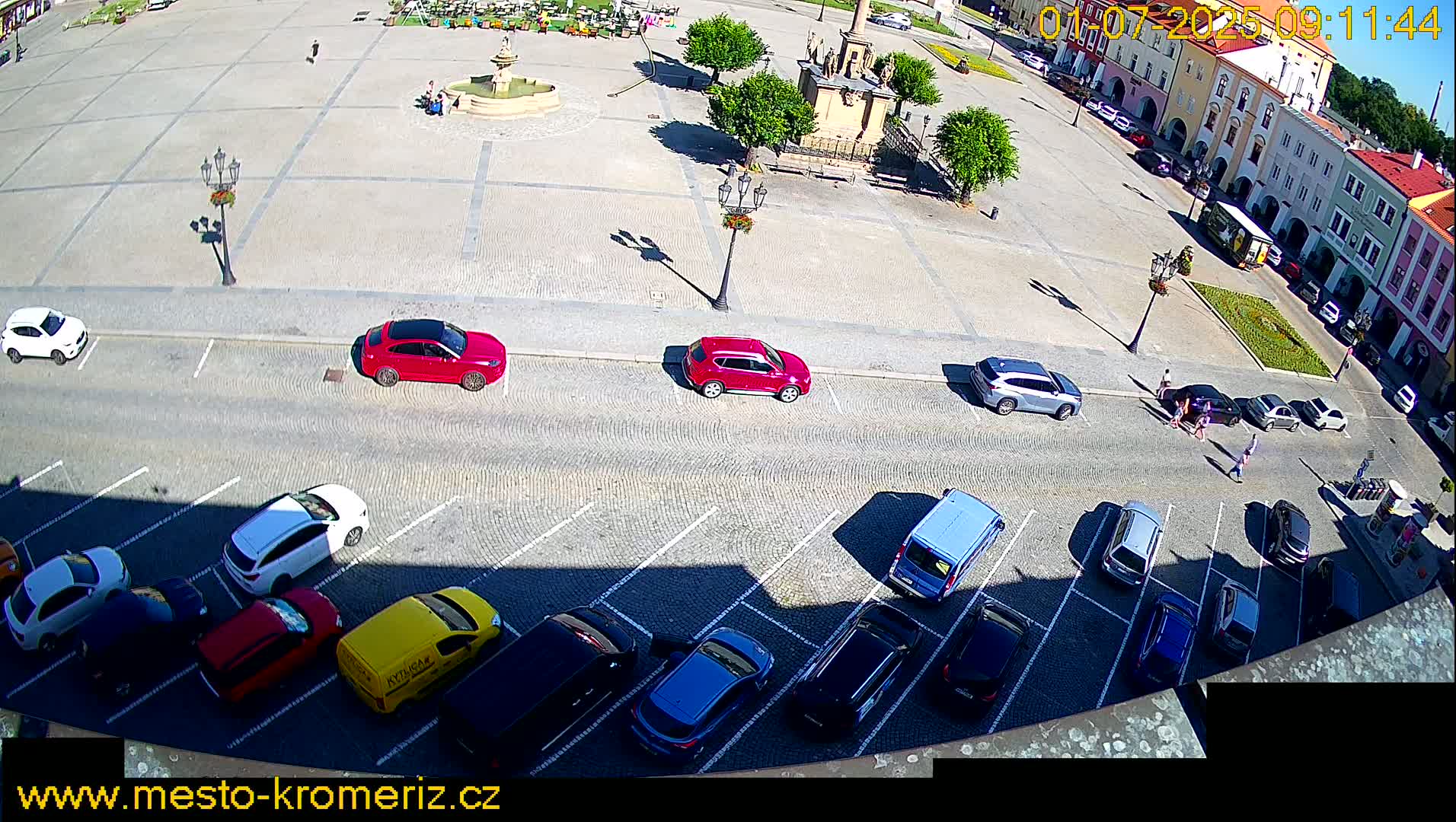 A sunny day shows a town square with a fountain and several cars parked in a lot adjacent to a street.