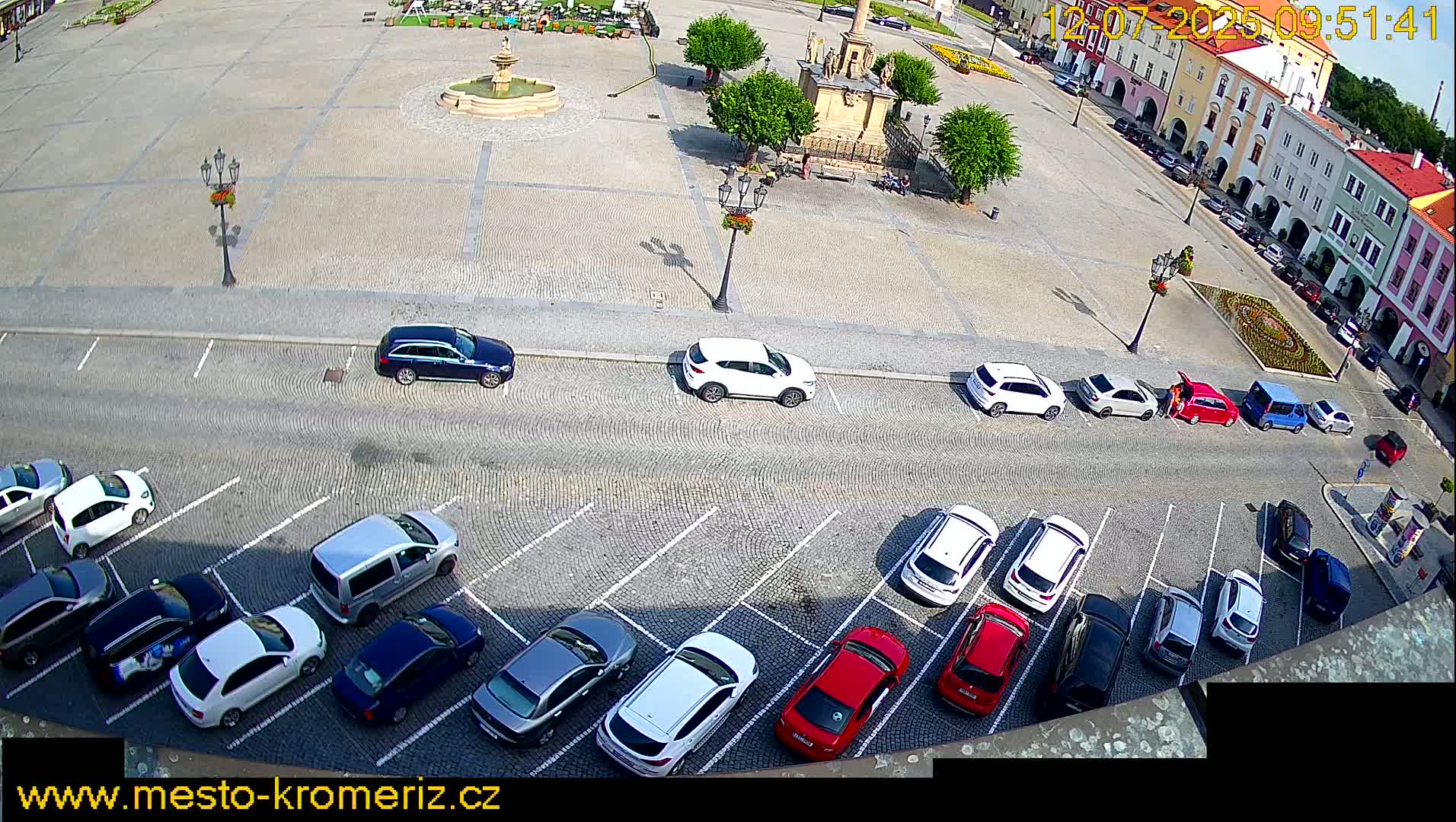 A sunny day in a cobblestone town square, with cars parked in rows along the perimeter and a fountain at the center.