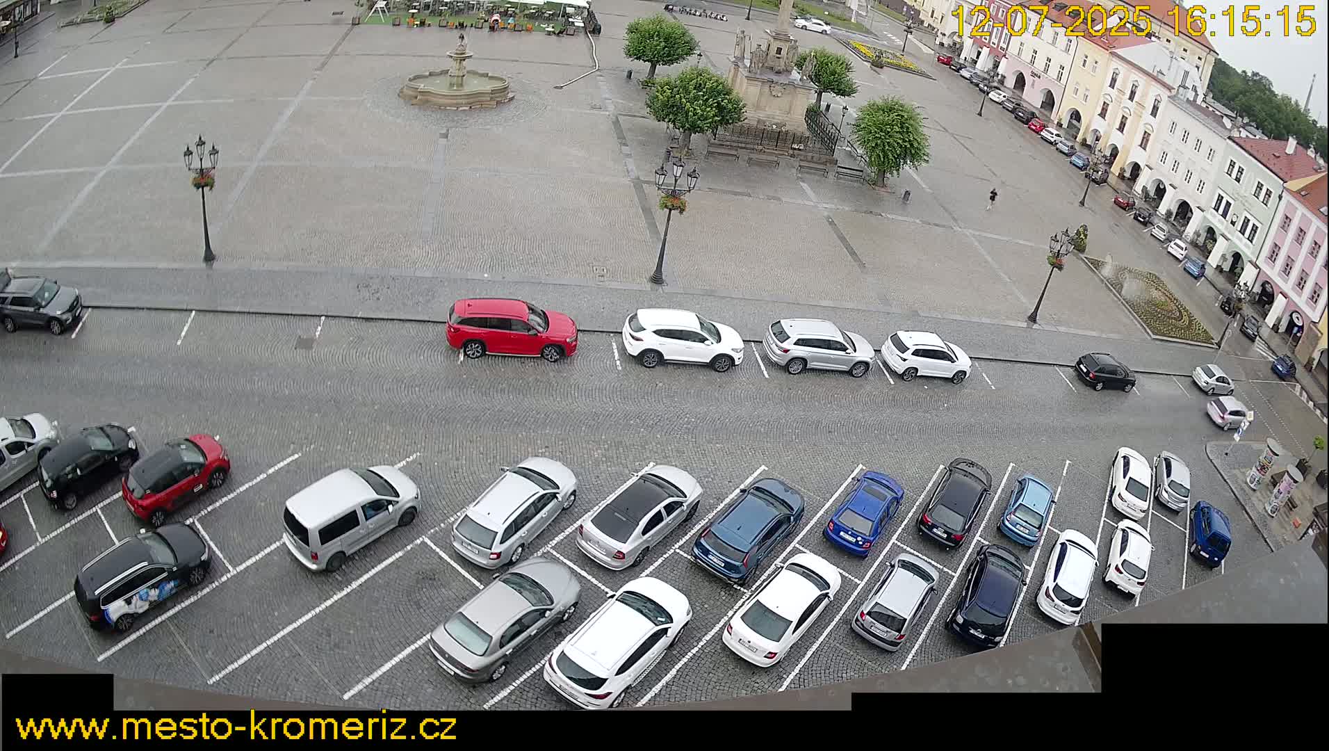 A sunny day in a cobblestone town square, with cars parked in rows along the perimeter and a fountain at the center.