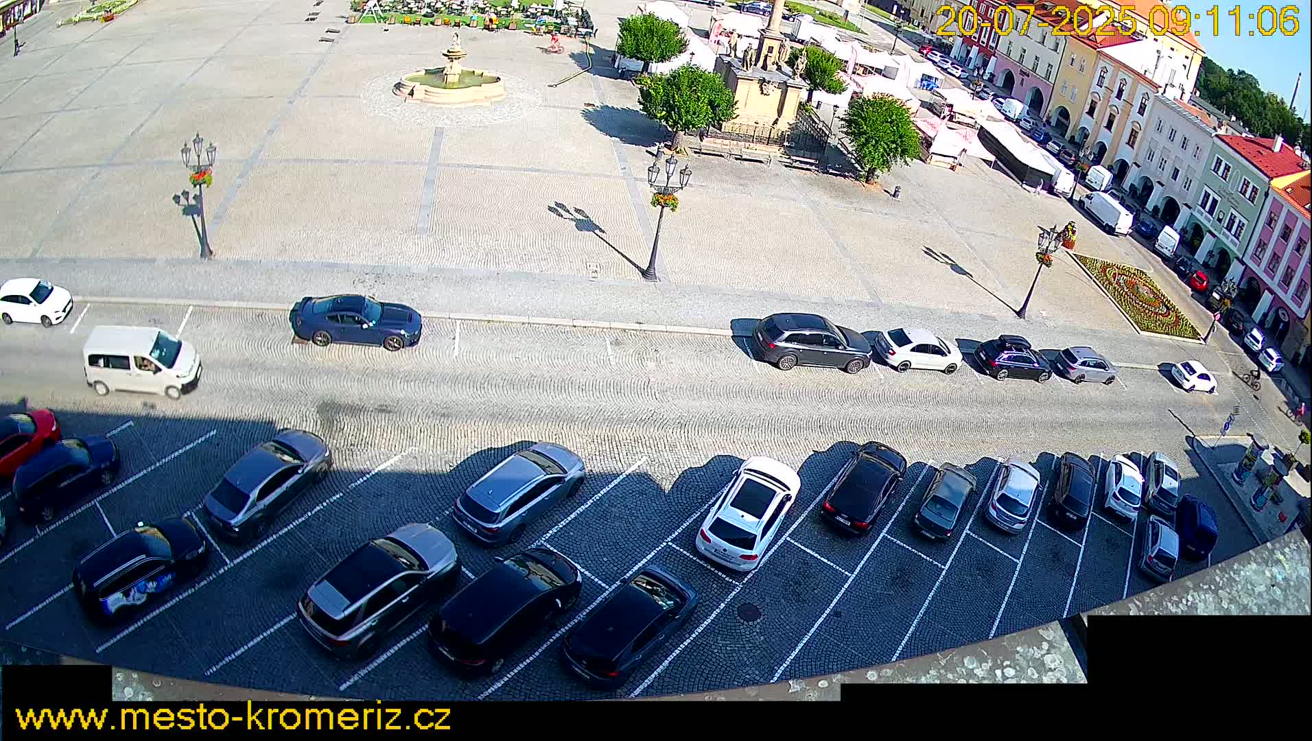 A town square with a fountain is surrounded by buildings and a parking lot filled with cars on a sunny day.