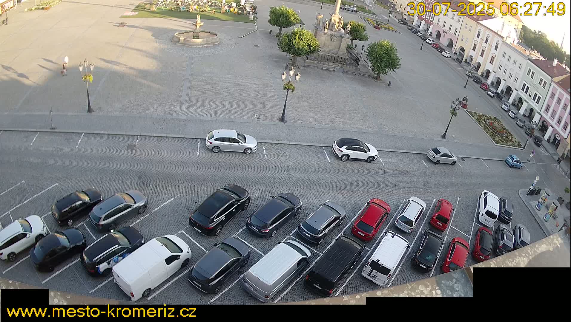 A town square with a fountain is surrounded by buildings and a parking lot filled with cars on a sunny day.