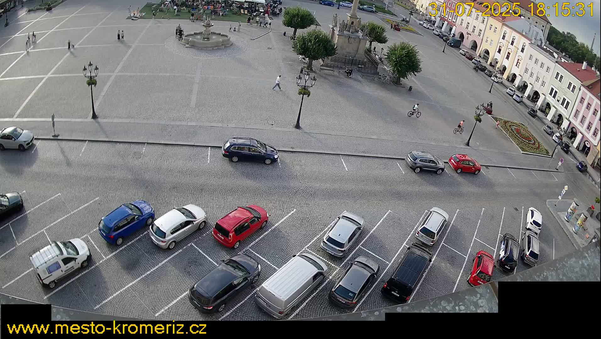 A paved town square with a fountain, some buildings, and parked cars, under a partly sunny sky.