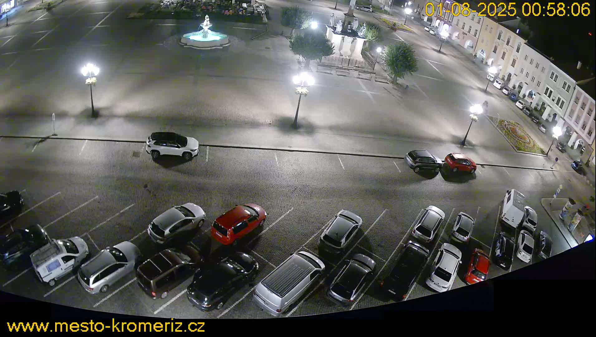 A nighttime overhead view of a town square's parking lot, filled with cars, and a fountain in the center of the square.