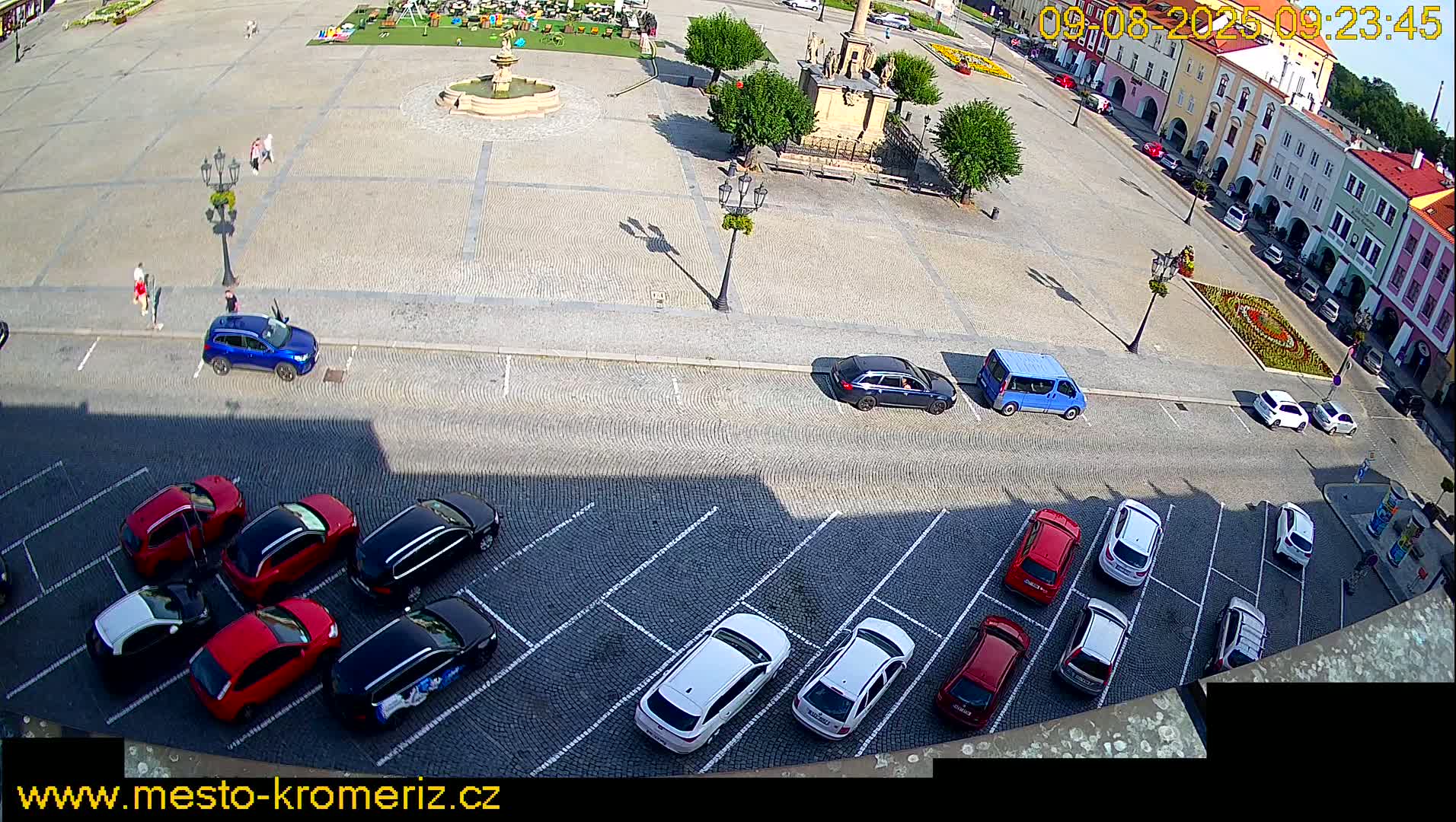 A sunny town square with a fountain, a monument, and several cars parked in a lot surrounding it.
