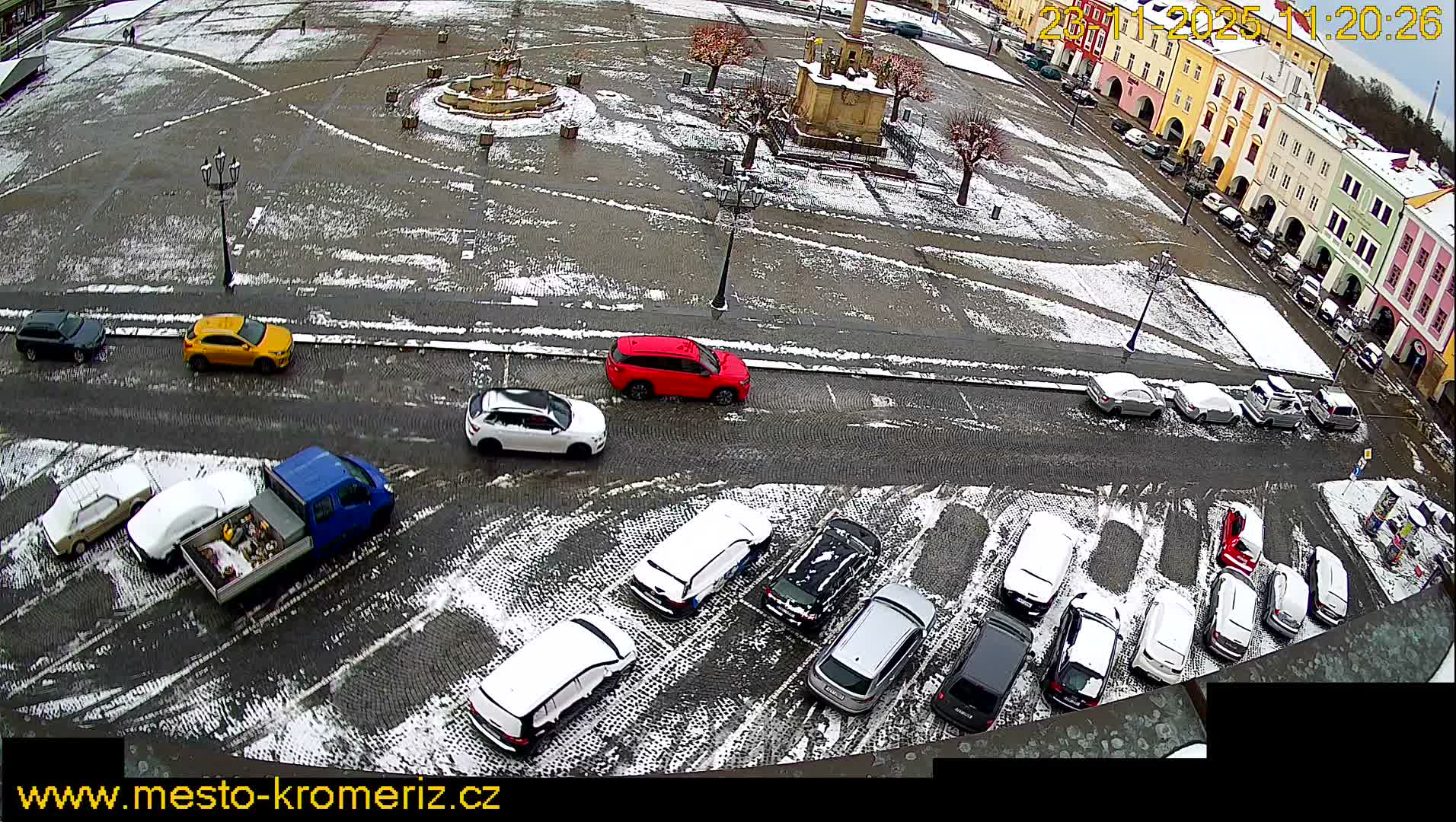 An aerial view reveals a snowy cobblestone town square with vehicles parked and driving, colorful buildings, and a central fountain under a bright winter sky.