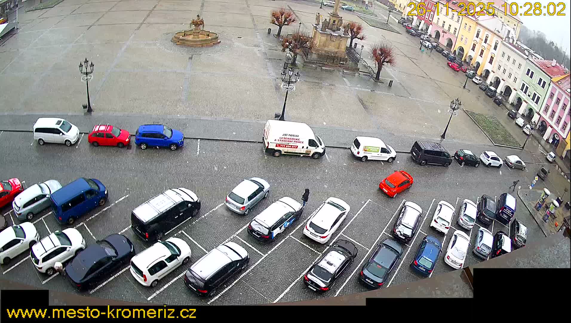 A wide, elevated view captures a wet, cobblestone town square bustling with numerous parked and a few moving cars, flanked by colorful historic buildings and monuments under an overcast sky with light precipitation.