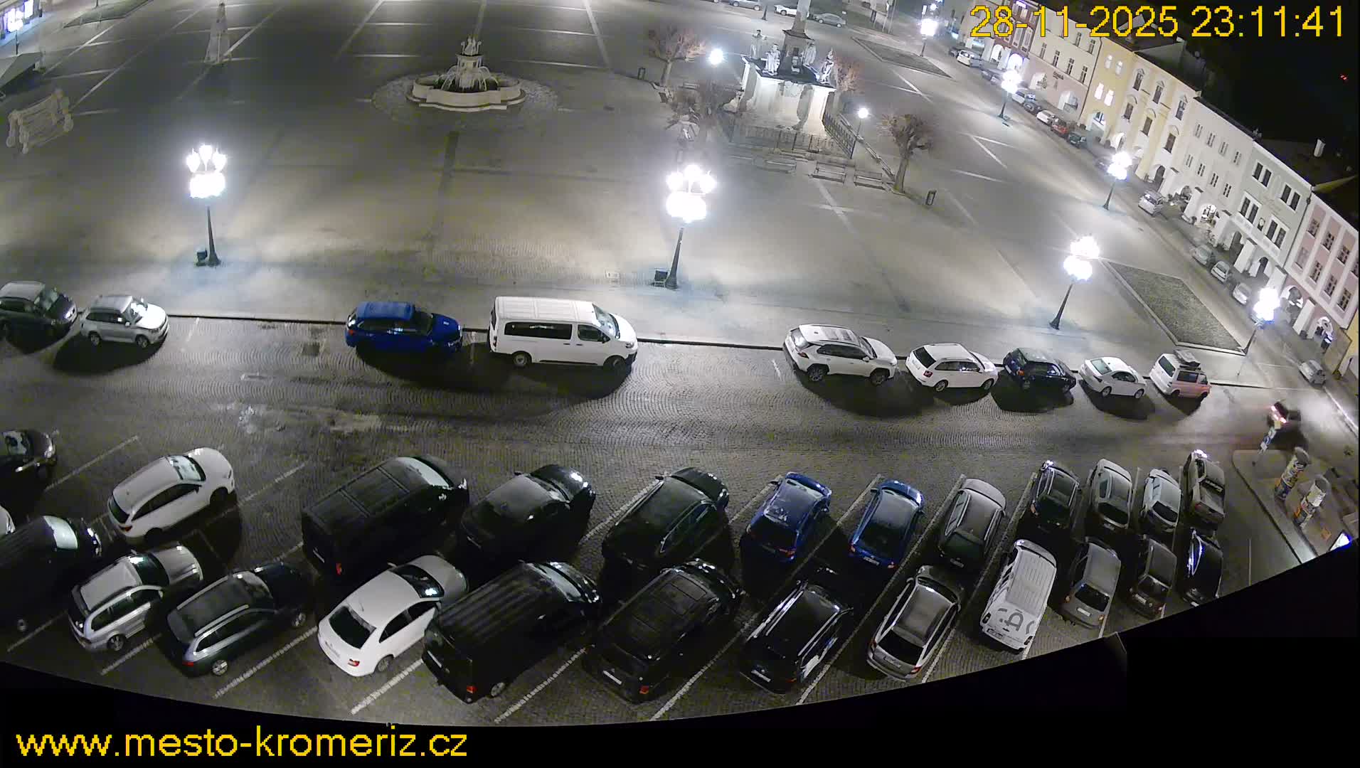 At night, a damp town square is illuminated by streetlights, featuring a fountain, surrounding buildings, and numerous parked cars.