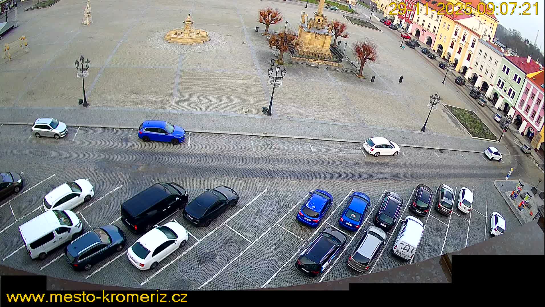 An overhead view reveals a cobbled town square filled with parked cars, featuring a central fountain and monument, surrounded by colorful buildings under an overcast sky.