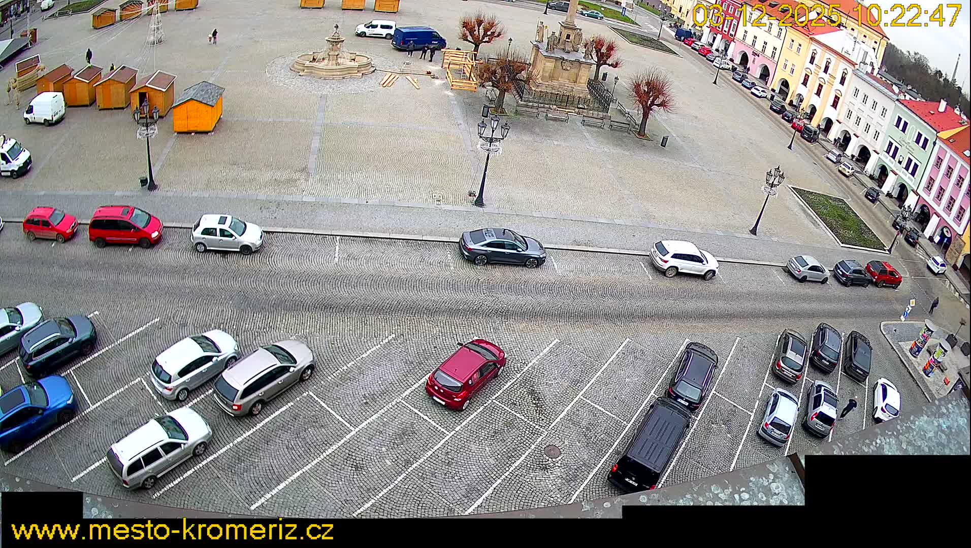 An overhead view captures a cobblestone town square on an overcast day, featuring wooden market stalls, bare trees, and architectural features, all bordered by a busy street filled with parked cars and a lively row of colorful, multi-story buildings.