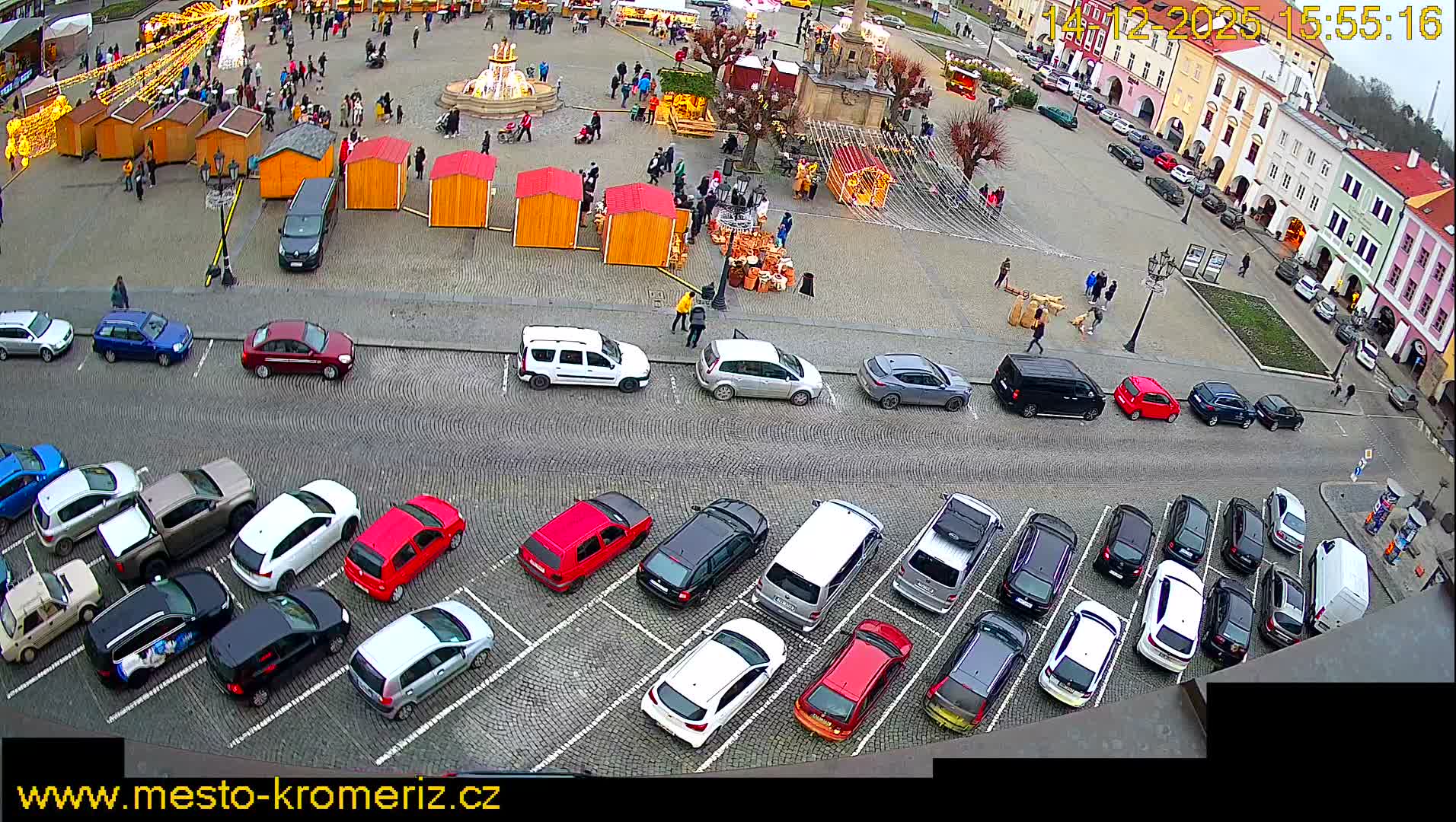 An overhead view captures a cobblestone town square on an overcast day, featuring wooden market stalls, bare trees, and architectural features, all bordered by a busy street filled with parked cars and a lively row of colorful, multi-story buildings.