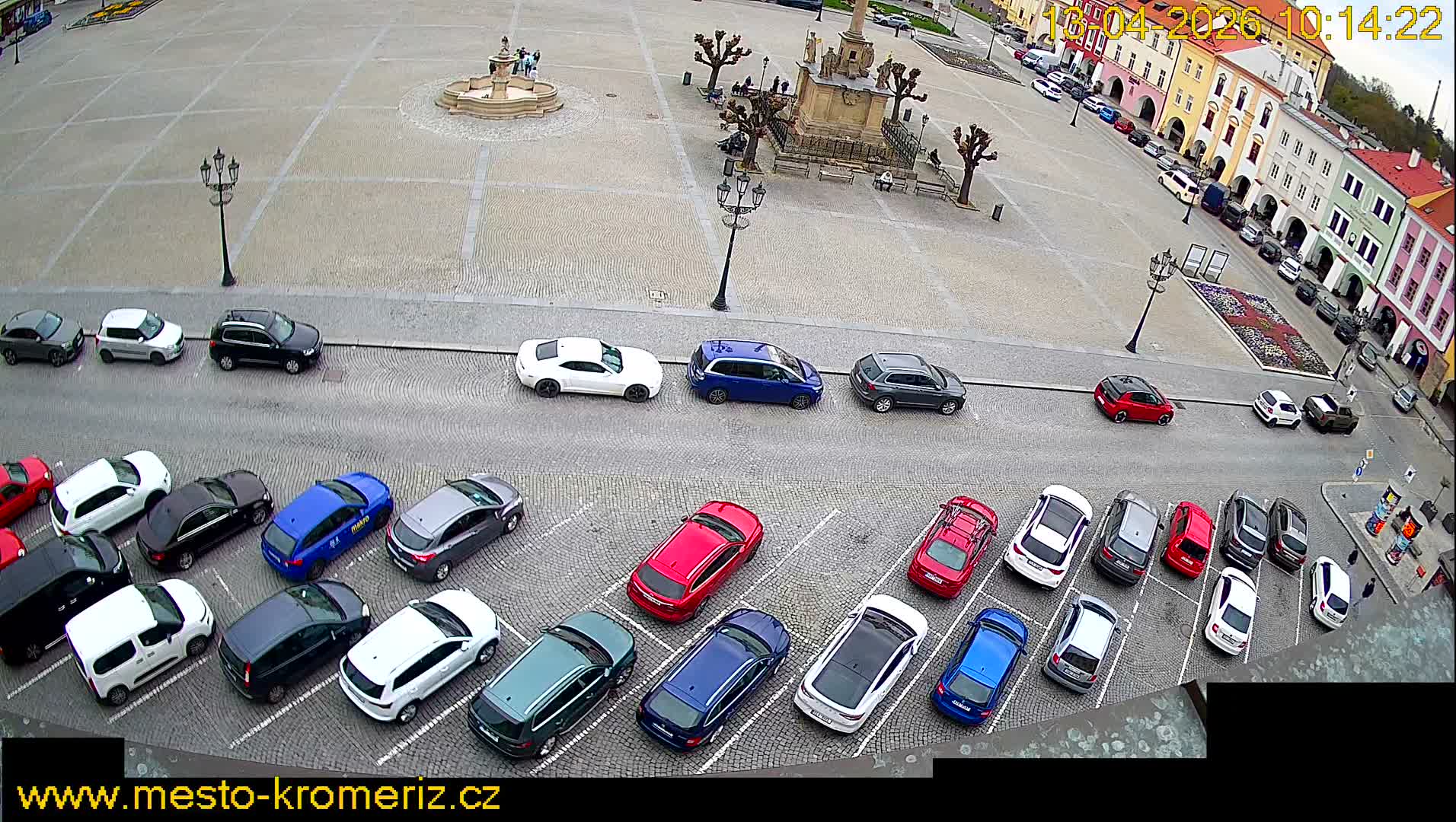 An overhead view captures a cobblestone town square on an overcast day, featuring wooden market stalls, bare trees, and architectural features, all bordered by a busy street filled with parked cars and a lively row of colorful, multi-story buildings.