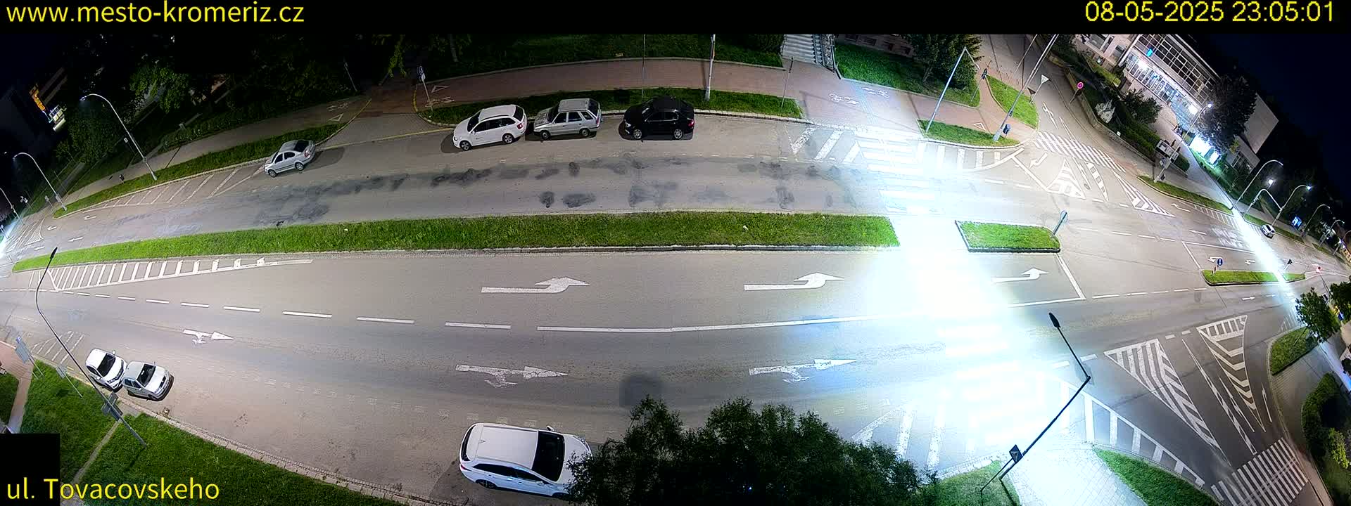 A high-angle nighttime view of a mostly empty road intersection with several parked cars and a few vehicles driving.