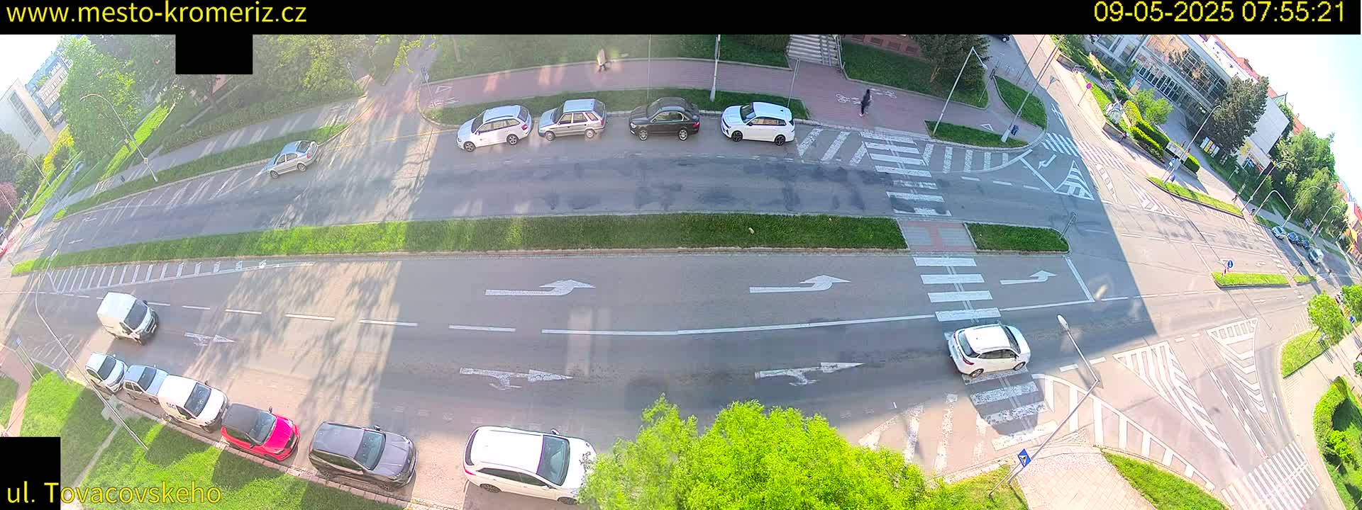 An aerial view of a sunny street intersection with several cars parked and driving, and pedestrians crossing.