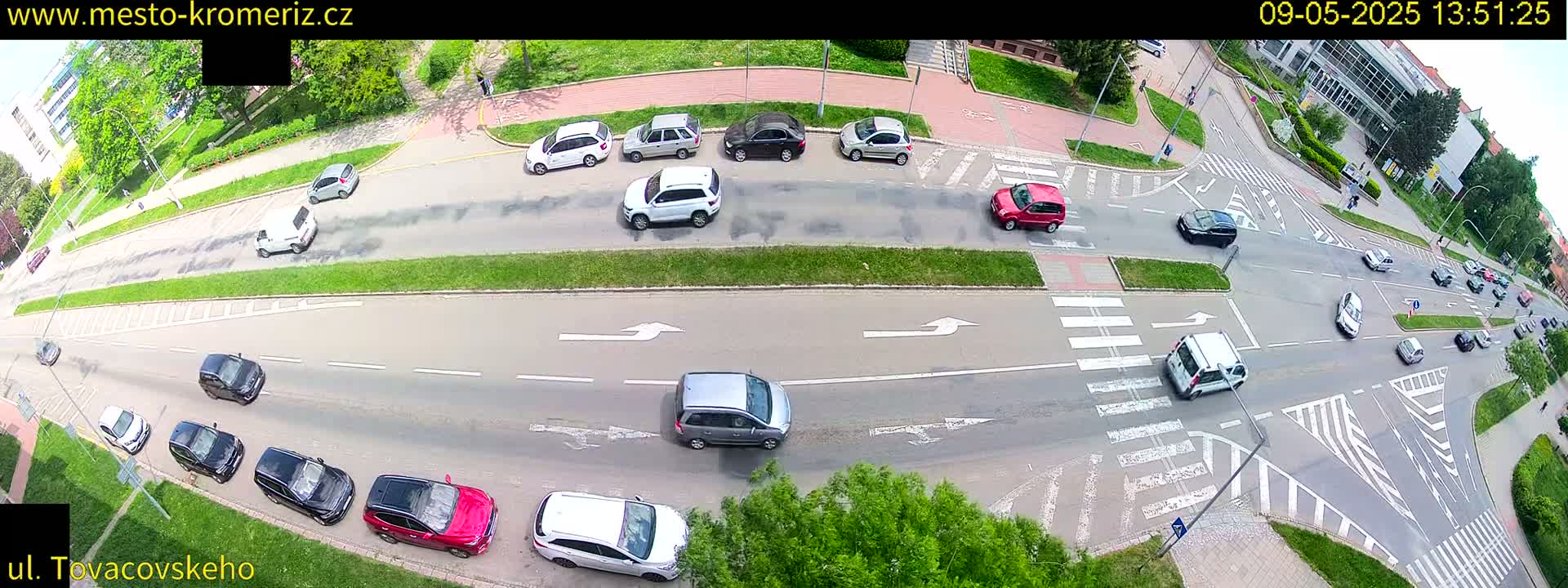 An aerial view of a road intersection with numerous cars, some parked and others moving, on a sunny day.