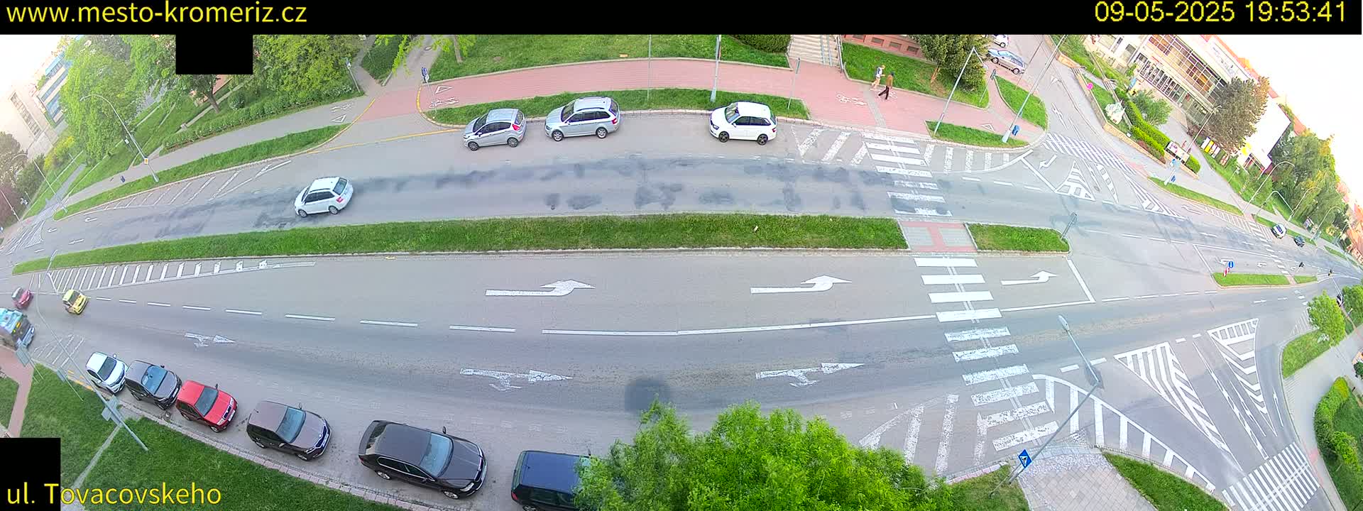 An aerial view of a street intersection with several parked cars and a few cars in motion, under partly sunny conditions.