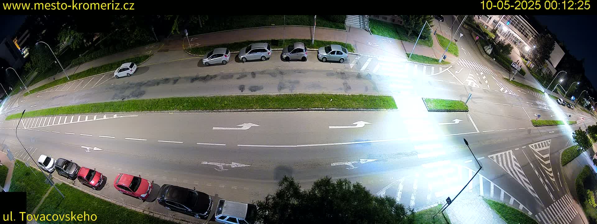 A nighttime aerial view shows a street intersection with parked cars along the sides and a few cars driving through the intersection under streetlights.