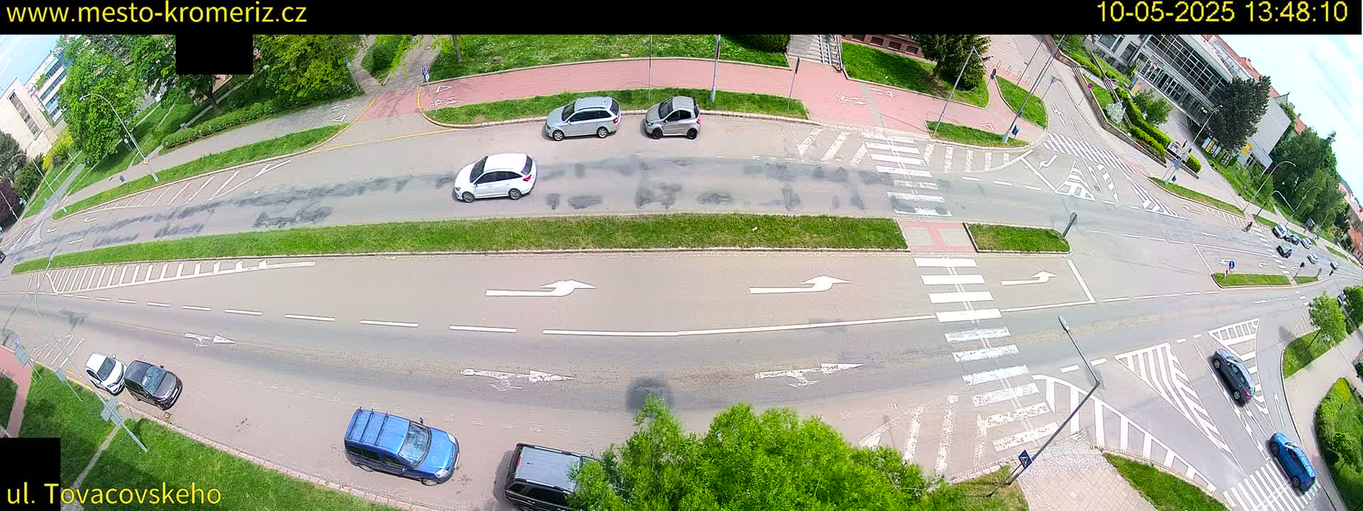 A high-angle view of a road intersection with several cars, pedestrians, and marked crosswalks on a sunny day.