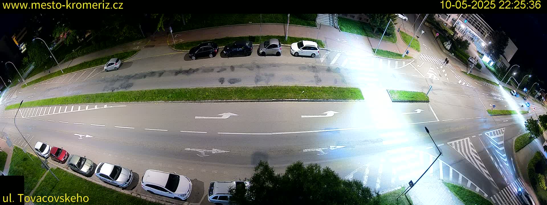 A nighttime, high-angle view of a street intersection with several parked cars and a few vehicles driving, showing street markings and surrounding greenery.