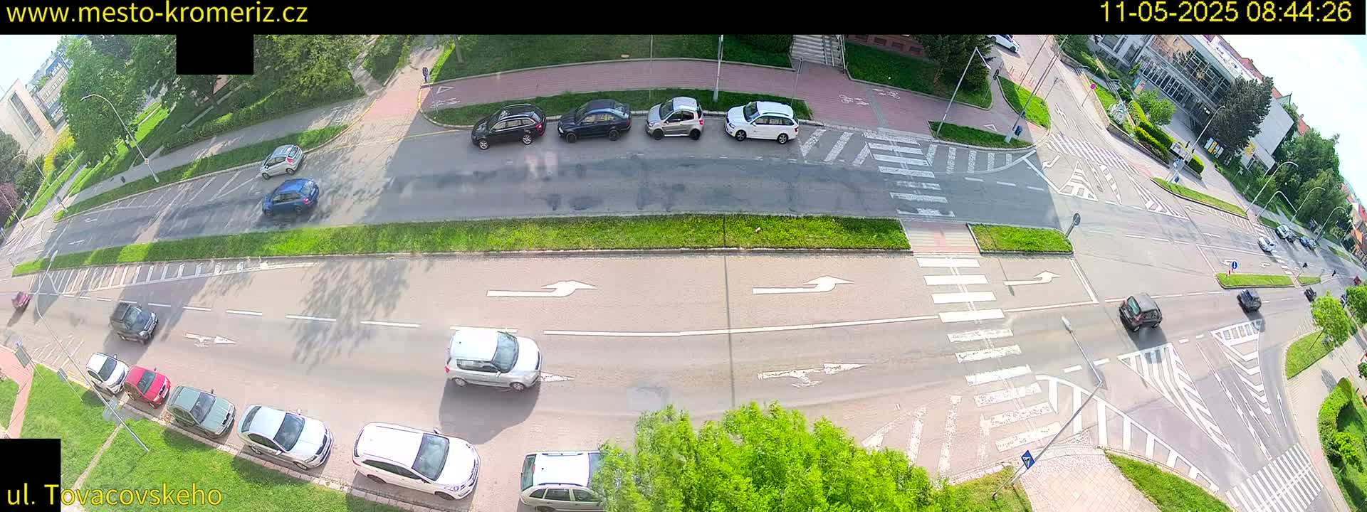 A sunny overhead view of a multi-lane road intersection with several cars driving and parked, surrounded by green grass and trees.