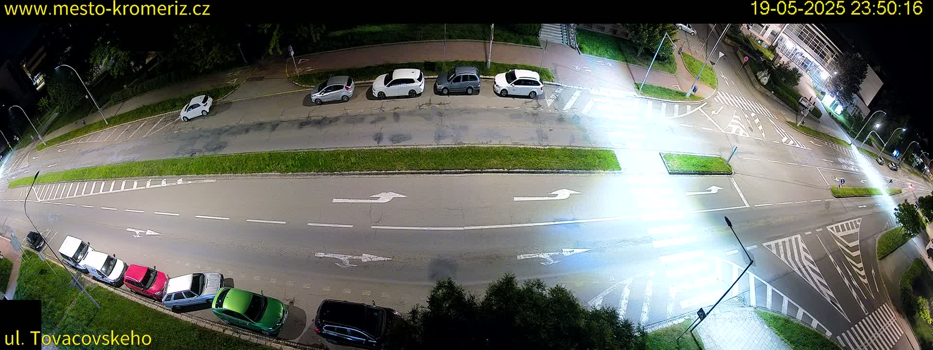 A nighttime overhead view of a street intersection with several cars parked along the side and a few cars driving on the road.
