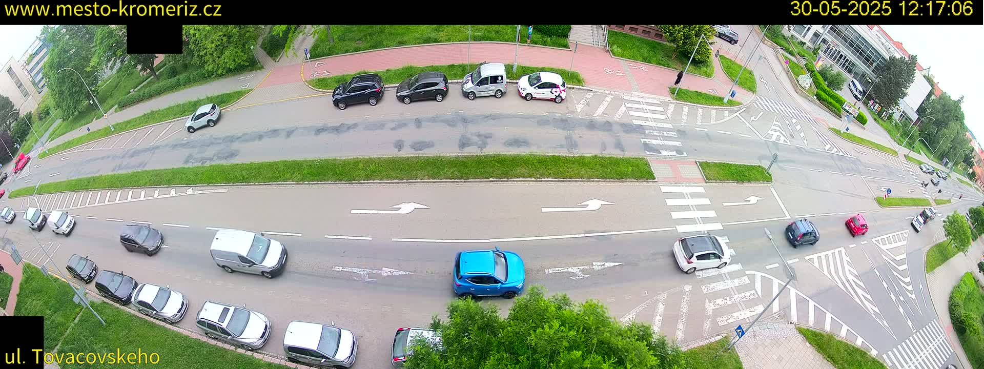 An aerial view of a road intersection with several cars and vans, surrounded by green landscaping under a partly cloudy sky.