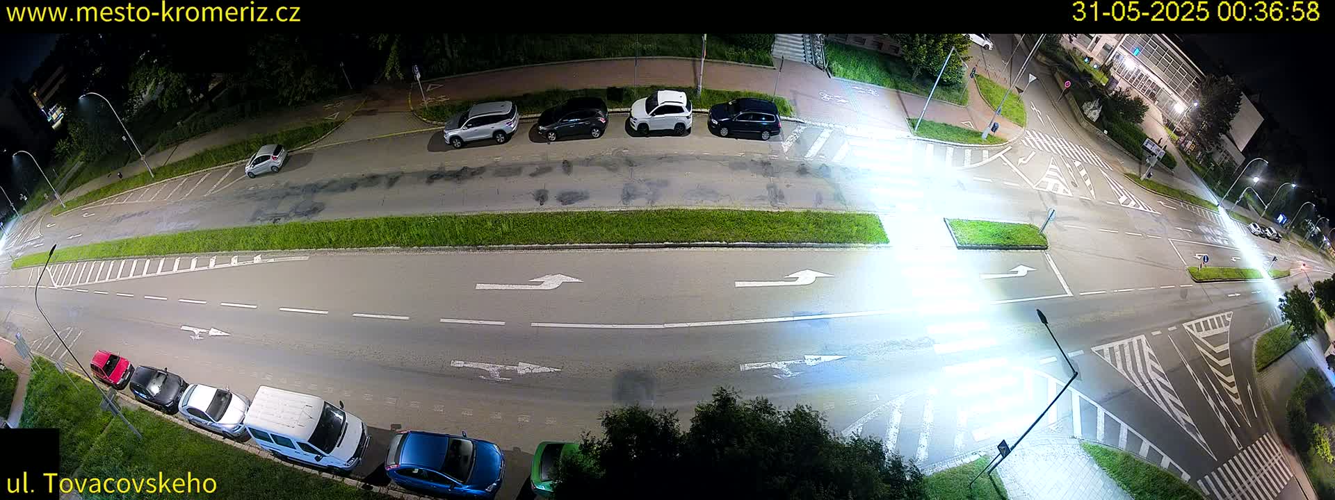 A nighttime, overhead view of a street intersection with several parked cars and few cars driving on the road, illuminated by streetlights.