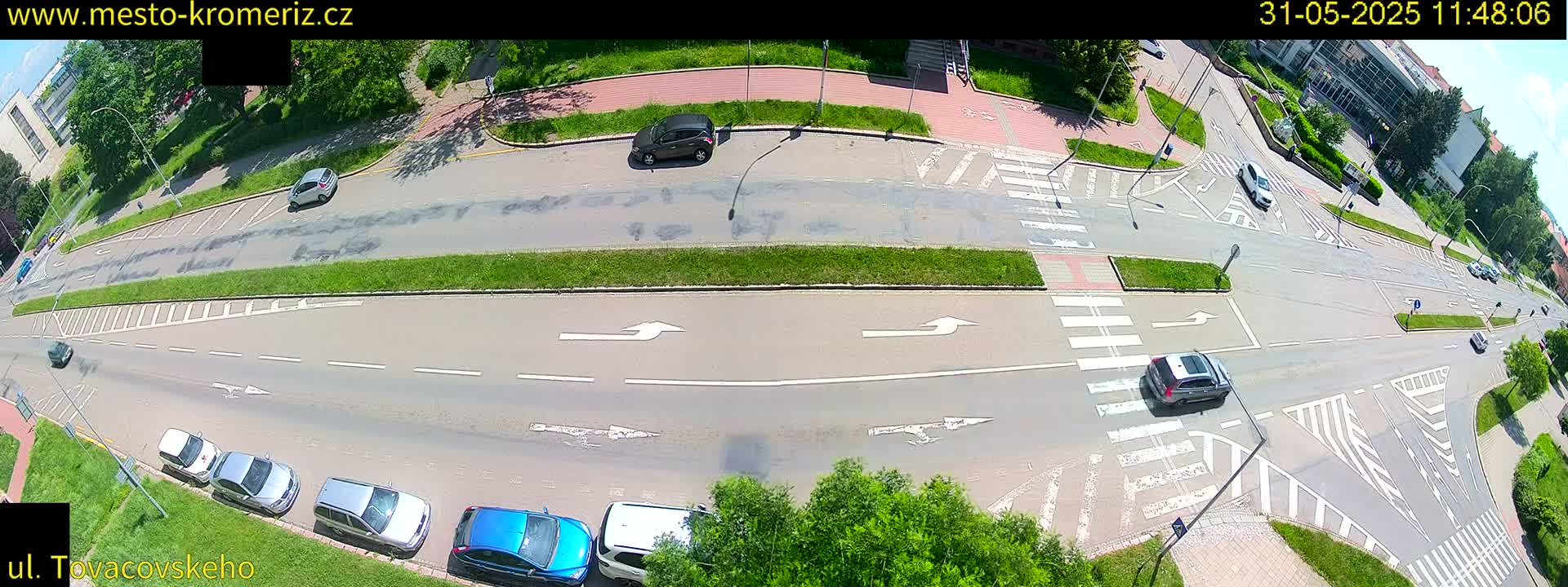 A high-angle view of a sunny day shows a multi-lane road intersection with several cars and green landscaping.