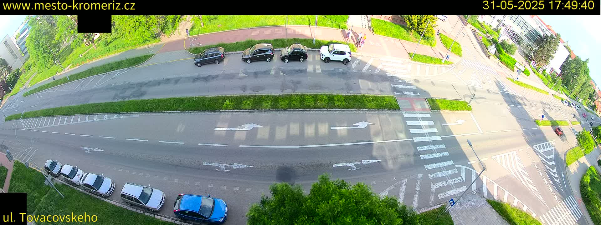 A sunny overhead view of a street intersection with several parked cars and a few cars driving, along with pedestrian crosswalks.