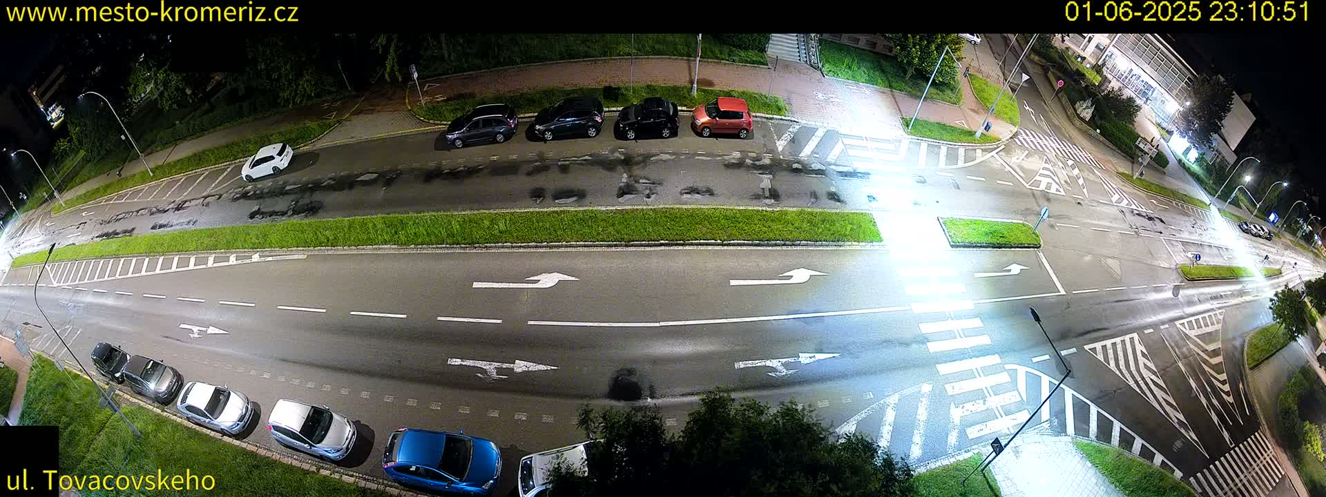 A nighttime, overhead view of a wet road intersection with cars parked along the sides and a few cars driving through.
