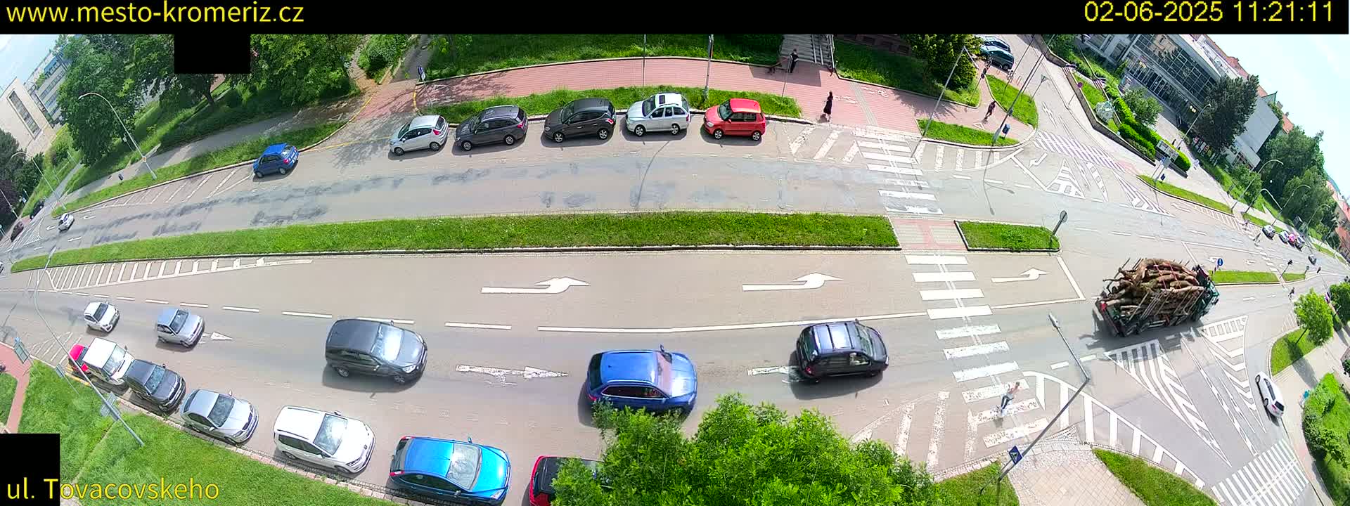 An aerial view of a city intersection on a sunny day shows several cars driving on a multi-lane road, with a truck carrying a load of logs turning at the intersection.
