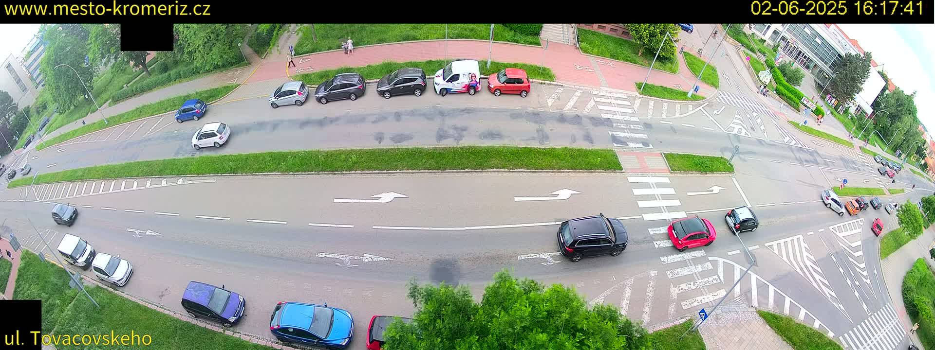 An overhead view shows several cars parked and driving on a multi-lane road and intersecting crosswalks on a partly cloudy day.