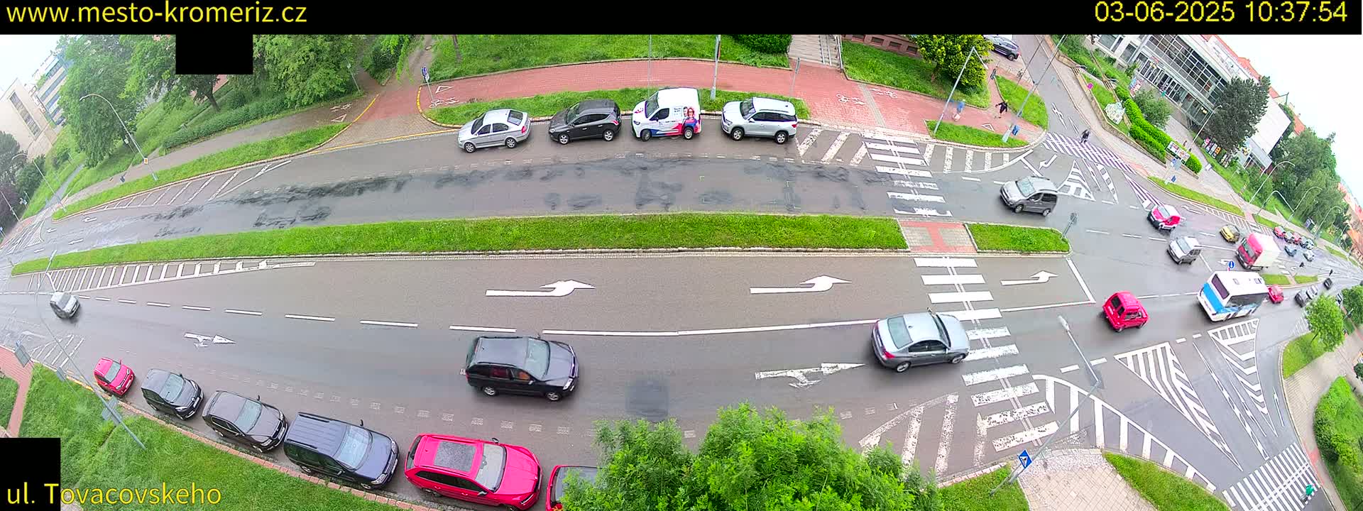 A high-angle view of a wet road intersection with several cars parked and driving under overcast skies.