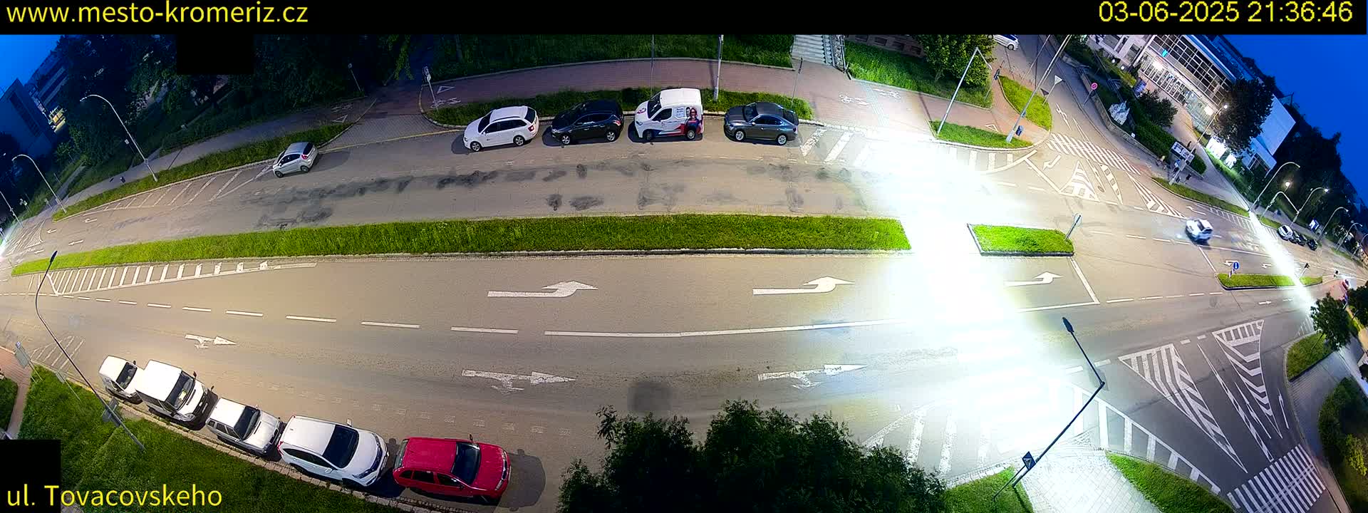 An aerial nighttime view of a street intersection with cars parked along the side and a few cars driving through.