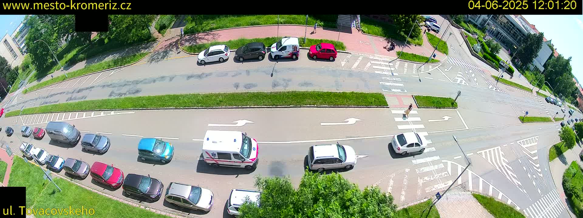 An overhead view of a sunny street intersection with numerous cars parked and driving, an ambulance, and a pedestrian crossing.