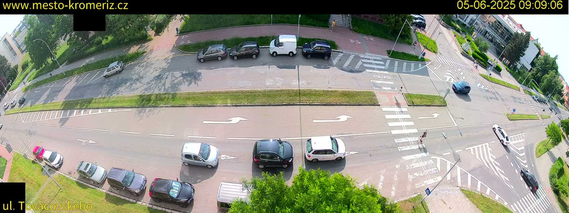 A sunny overhead view of a multi-lane road intersection with several cars, parked and moving, and pedestrians crossing a crosswalk.