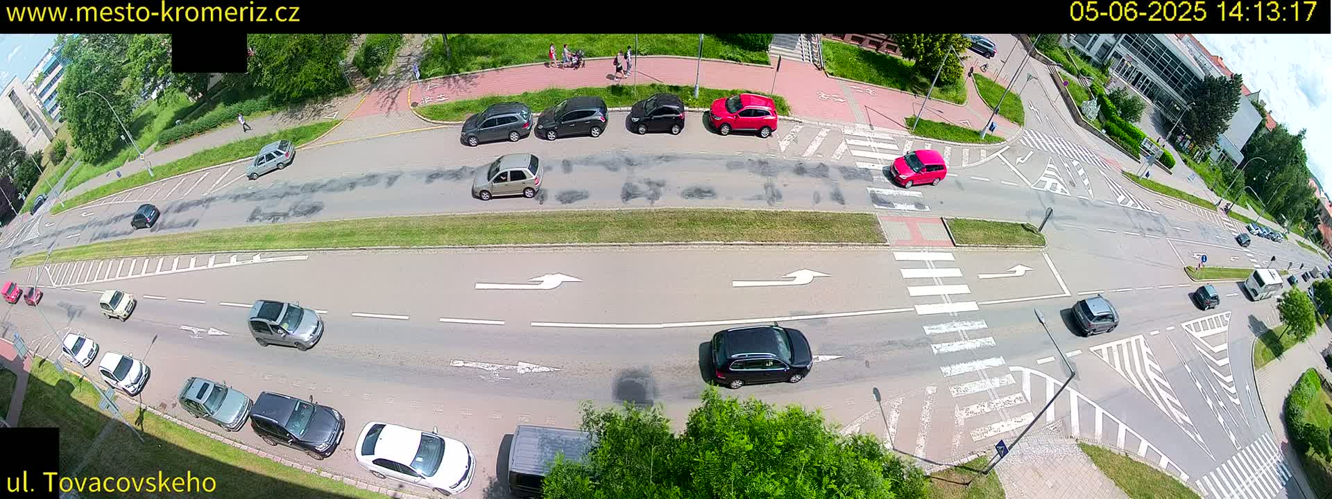 An aerial view of a sunny day shows a multi-lane road intersection with numerous cars and pedestrians, marked by crosswalks and directional arrows.
