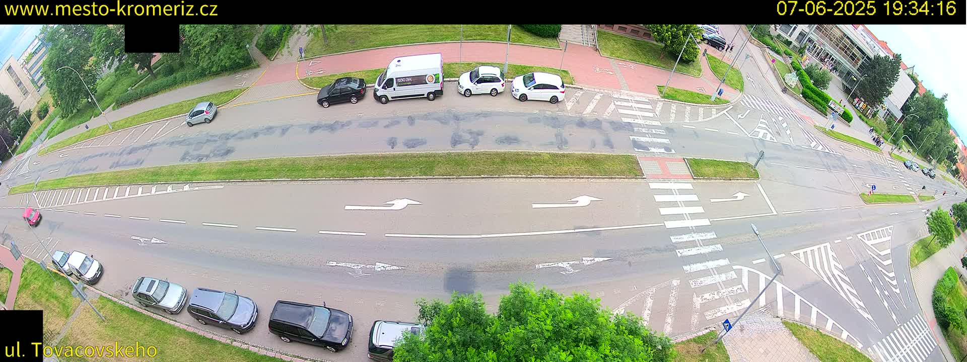 A high-angle view of a street intersection with several cars parked and driving, under a mostly clear sky.