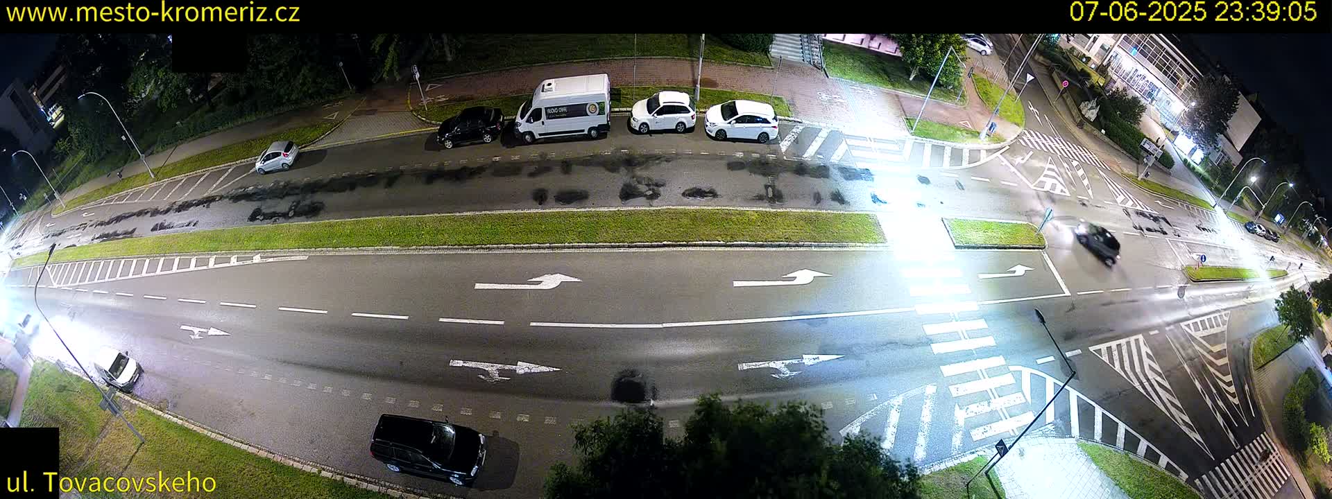 A nighttime high-angle view of a multi-lane road intersection with several cars, a van, and pedestrian crosswalks, wet from recent rain.