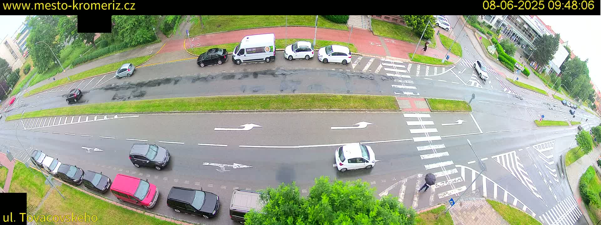 A wet, overhead view of a street intersection shows several cars parked and driving, with a pedestrian crossing under an umbrella.