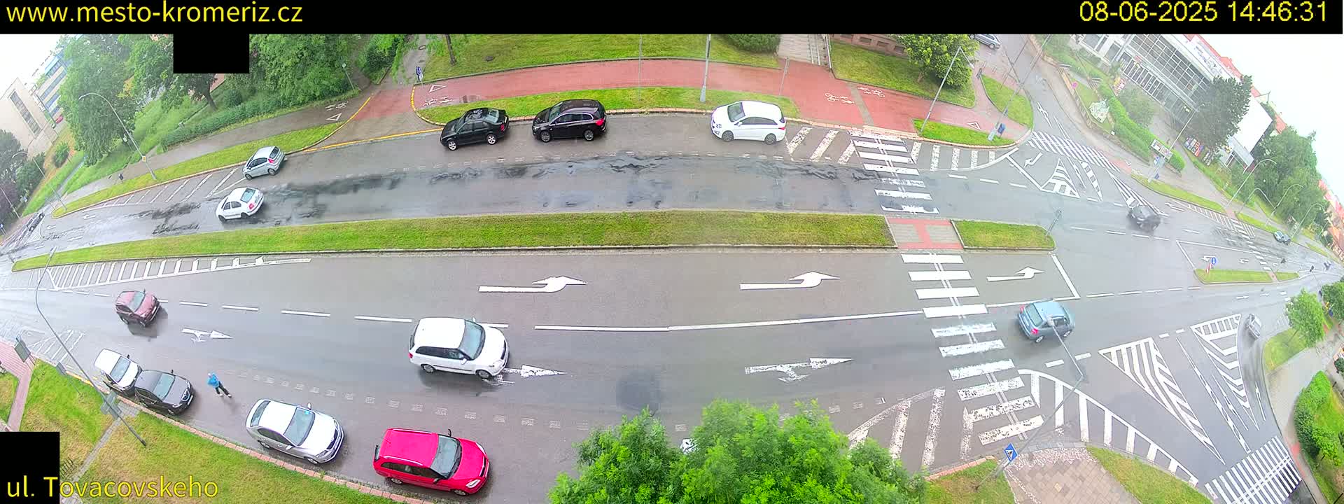A wet road intersection with several cars and a pedestrian, under a cloudy sky.